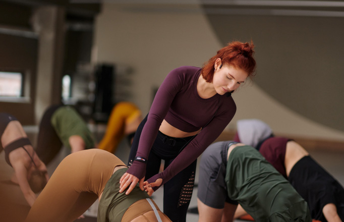 Bild: Eine Kursleiterin korrigiert bei einer Teilnehmenden die Haltung in der Yoga-Pose „Herabschauender Hund“. Weitere Personen sind im Hintergrund zu sehen.
