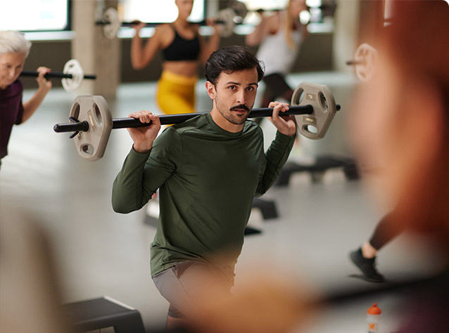 Bild: Ein Mann macht im Kursraum mit einer Langhantel auf dem Rücken einen Ausfallschritt. Weitere Teilnehmende führen dieselbe Übung aus.