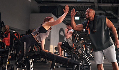 Bild: Eine Frau und ein Mann geben sich nach einer Trainingseinheit im Freihantelbereich ein High Five. Beide lächeln sich motivierend an.