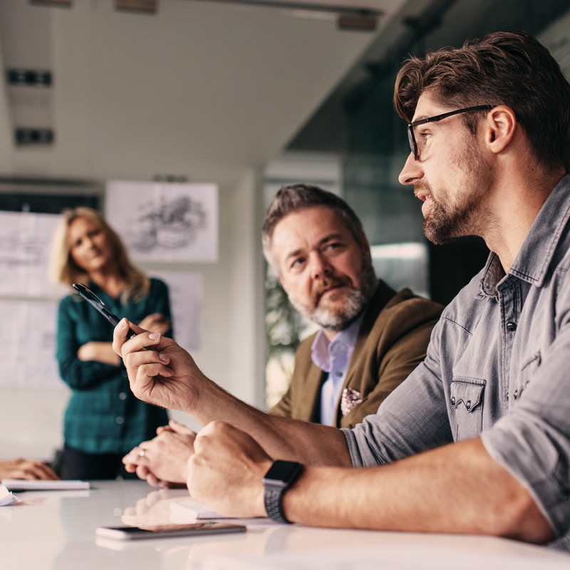 Bild: Mehrere Personen sitzen an einem Konferenztisch. Ein Mann im Vordergrund gestikuliert mit einem Stift. Im Hintergrund steht eine Frau vor ausgedruckten Unterlagen.