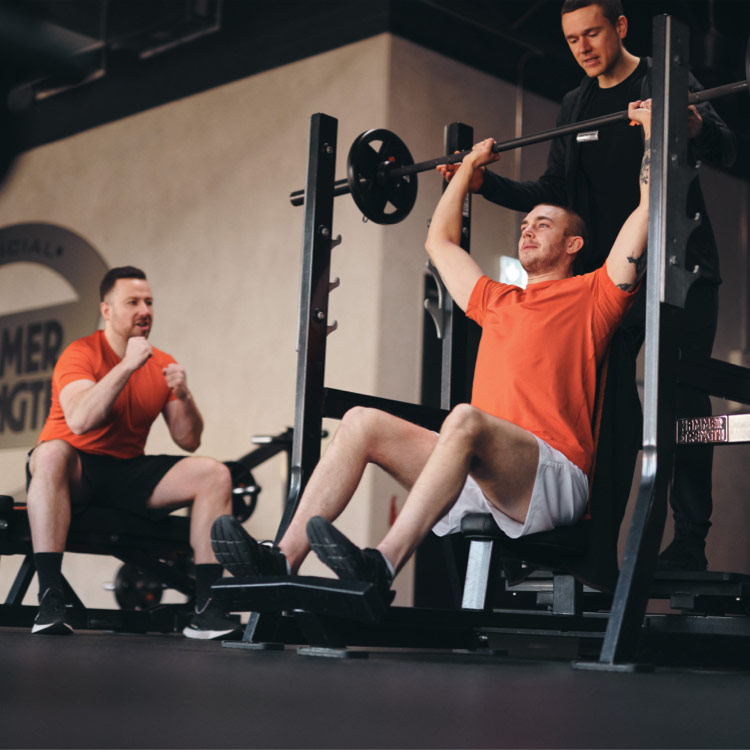 Bild: Ein Mann führt unter Anleitung eines FitX-Trainers Schulterpressen mit Langhanteln aus. Ein weiterer Trainierender beobachtet ihn und feuert ihn an.