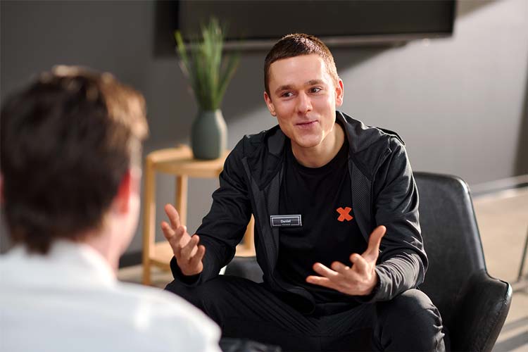 Bild: Ein FitX-Mitarbeiter spricht in einem Beratungsgespräch mit einer weiteren Person. Er lächelt und gestikuliert freundlich mit den Händen.